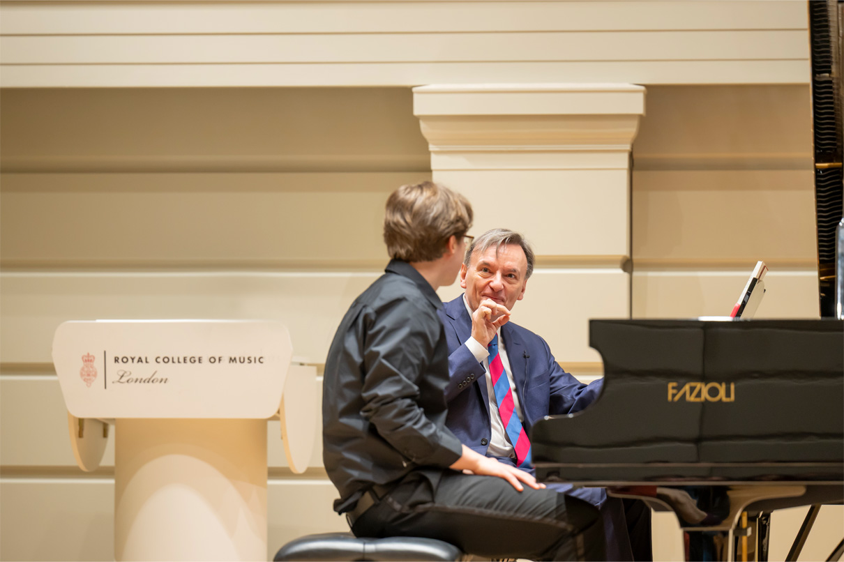 Two men, one a student, one a teacher, instructing a piano performance on a stage, with other students listening.